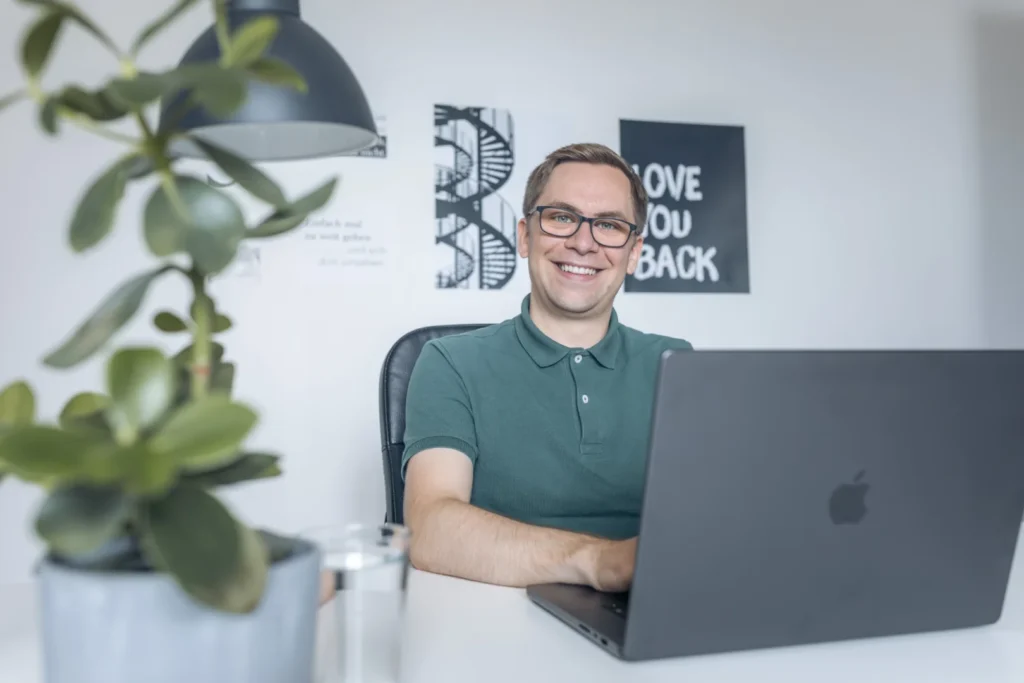 Photo of Nicolas Wazulek sitting at his MacBook and smiling into the camera. Green plant blurred on the left of the picture. In the background are pictures hanging on the wall.