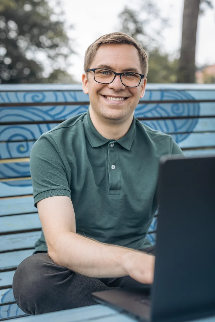 Nicolas Wazulek working outside on a big blue bench smiling into the camera.